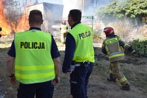 Umundurowani policjanci stoją na terenie posesji. W oddali palą się gałęzie. Po prawej stronie biegnie strażak z linią gaśniczą