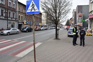 Umundurowana policjantka oraz policjant stoją na chodniku przy oznakowanym przejściu dla pieszych. W tle oznakowany radiowóz
