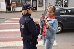 Umundurowana policjantka stoi z kobietą na chodniku przy oznakowanym przejściu dla pieszych i wręcza jej ulotkę informacyjną