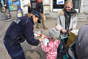 Umundurowana policjantka wręcza dziecku opaskę odblaskową. Z boku stoi kobieta z wózkiem