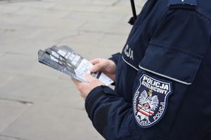 Zbliżenie na naszywkę znajdującą się na ,mundurze policjanta &quot;Policja dzielnicowy Komenda Miejska Policji&quot;. Policjant trzyma w rękach ulotki informacyjne
