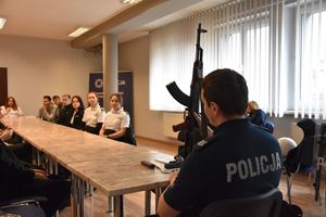 Umundurowany policjant siedzi przy stole i pokazuje uczniom broń