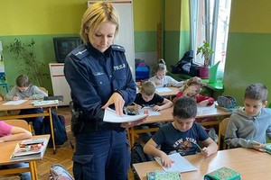 Umundurowana policjantka podczas spotkania z dziećmi w szkole podstawowej rozdaje materiały profilaktyczne W ławkach siedzą dzieci.