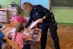 Umundurowana policjantka podczas spotkania z dziećmi w szkole podstawowej rozdaje materiały profilaktyczne W ławkach siedzą dzieci.