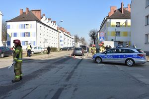 Miejsce zdarzenia drogowego. Strażak reguluje ruchem na środku skrzyżowania. Z boku stoi zaparkowany oznakowany policyjny radiowóz. W tle policjant wykonują czynności procesowe