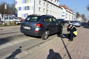 Miejsce zdarzenia drogowego. Za oznakowanym przejściem dla pieszych stoi pojazd osobowy oraz policjant, który wykonuje czynności procesowe. W tle zaparkowany pojazdy oraz budynki mieszkalne