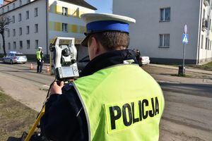 Umundurowany policjant w kamizelce odblaskowej z napisem &quot;Policja&quot; obsługuje tachimetr w miejscy zdarzenia drogowego. W tle policyjny radiowóz oraz budynki mieszkalne