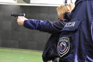 Zbliżenie na naszywkę &quot;Instruktor strzelań policyjnych&quot; znajdującą się na mundurze policjanta. W tle policjantka, która trzyma w rękach broń