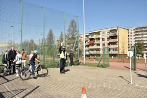 Miasteczko rowerowe. Umundurowany policjant ubrany w kamizelkę odblaskową stoi z ręką podniesioną do góry. Chłopiec na rowerze stoi obok policjanta