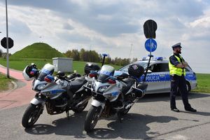 Umundurowany policjant stoi obok oznakowanego policyjnego radiowozu oraz motocykli. W tle Kopiec Wyzwolenia