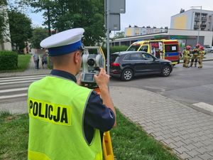 Policjant ruchu drogowego podczas obsługi wypadku drogowego. W tle pojazd osobowy