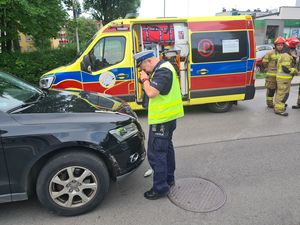 Policjant wykonujący zdjęcia pojazdu biorącego udział w zdarzeniu drogowym. W tle karetka pogotowia