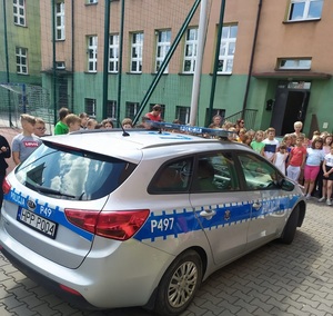 Policyjny radiowóz stoi przed budynkiem szkoły. Obok pojazdu stoją dzieci.