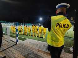 Policjant ubrany w kamizelkę odblaskowa. Z tyłu młodzi piłkarze