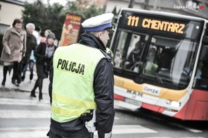 Na zdjęciu widać umundurowanego policjanta, który stoi na przejściu dla pieszych. W tle widać autobus z napisem cmentarz