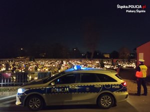 Policjant stoi obok radiowozu. W tle oświetlony cmentarz