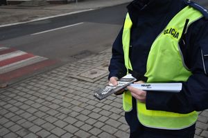 Policjant stoi przy oznakowanym przejściu dla pieszych. Mundurowy trzyma w ręku odblaski i ulotki