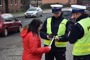 Policjanci ubrani w odblaskowe odblaski wręczają kobiecie odblask i ulotkę