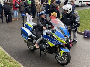 Zdjęcie przedstawia dziewczynę siedzącą na policyjnym motocyklu. Z boku umundurowany policjant