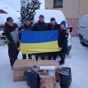 Zdjęcie przedstawia osoby trzymające ukraińską flagę. Na ziemi poukładane są dary