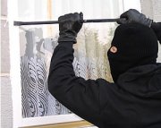 Zdjęcie przedstawia złodzieja ubranego na czarno z kominiarką na głowie, który próbuje wywarzyć okna za pomocą łomu.