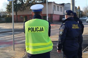 Zdjęcie przedstawia policjanta oraz strażników miejskich stojących przy oznakowanym przejściu dla pieszych