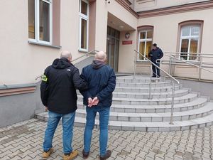 Zdjęcie przedstawia policjanta, który stoi z mężczyzną przed budynkiem prokuratury rejonowej