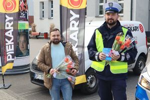 Zdjęcie przedstawia policjanta oraz prezentera Radia Piekary, którzy stoją obok radiowozu i trzymają w rękach kwiaty