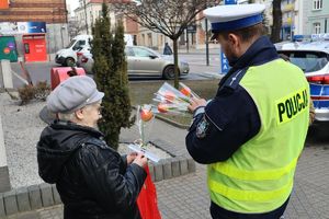 Zdjęcie przedstawia policjanta wręczającego kobiecie kwiatek oraz ulotkę informacyjną i odblask