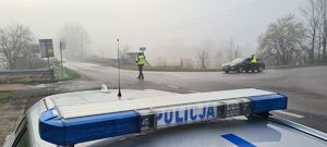 Zdjęcie przedstawia napis Policja umieszczony na dachu radiowozu. W tle policjanci ruchu drogowego