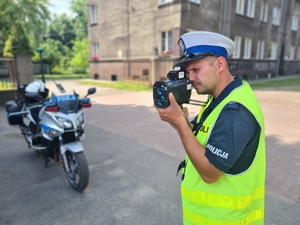 Zdjęcie przedstawia policjanta podczas kontroli prędkości. W tle policyjny motocykl