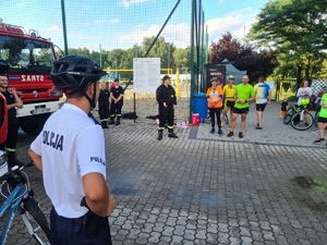 Zdjęcie przedstawia policjanta oraz uczestników wydarzenia. W tle wozy strażackie