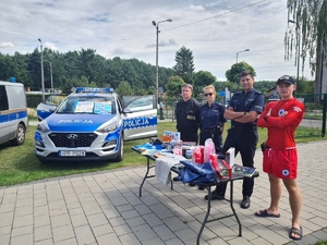 Zdjęcie przedstawia policjantów oraz strażaków i ratowników przy stoliku w tle radiowóz