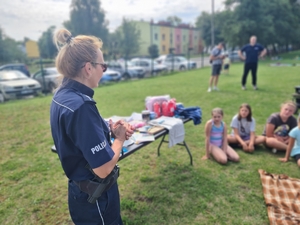 Zdjęcie przedstawia policjantkę, w tle dzieci siedzące na trawie