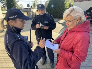 Zdjęcie przedstawia policjantów wręczających kobiecie ulotki i opaski odblaskowe