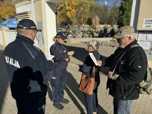 Zdjęcie przedstawia policjantów wręczających ulotki osobom przy cmentarzu