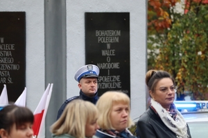 Zdjęcie przedstawia policjanta stojącego obok radiowozu na tle pomnika