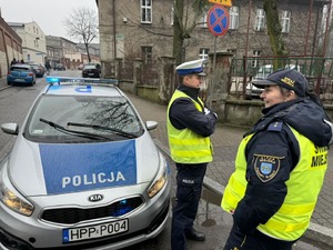 Zdjęcie przedstawia policjanta oraz strażniczkę miejską stojących przy radiowozie policyjnym