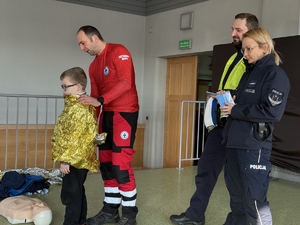 Zdjęcie przedstawia służby ratownicze podczas pogadanki oraz chłopca owiniętego w folię ratowniczą