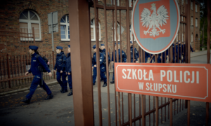 Zdjęcie bramy wejściowej do Szkoły Policji w Słupsku. W tle maszerujący policjanci