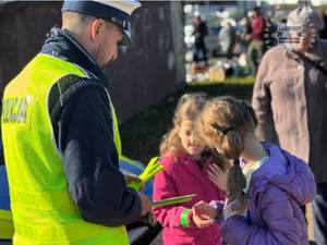 Zdjęcie przedstawia policjanta wręczającego przechodniom odblaskowe opaski