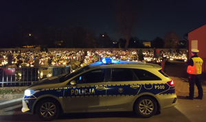 Zdjęcie policjanta stojącego przy radiowozie. W tle cmentarz