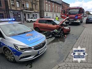 Policyjny radiowóz oraz motocykl i samochód osobowy biorący udział w zdarzeniu drogowym. W tle wóz Straży Pożarnej